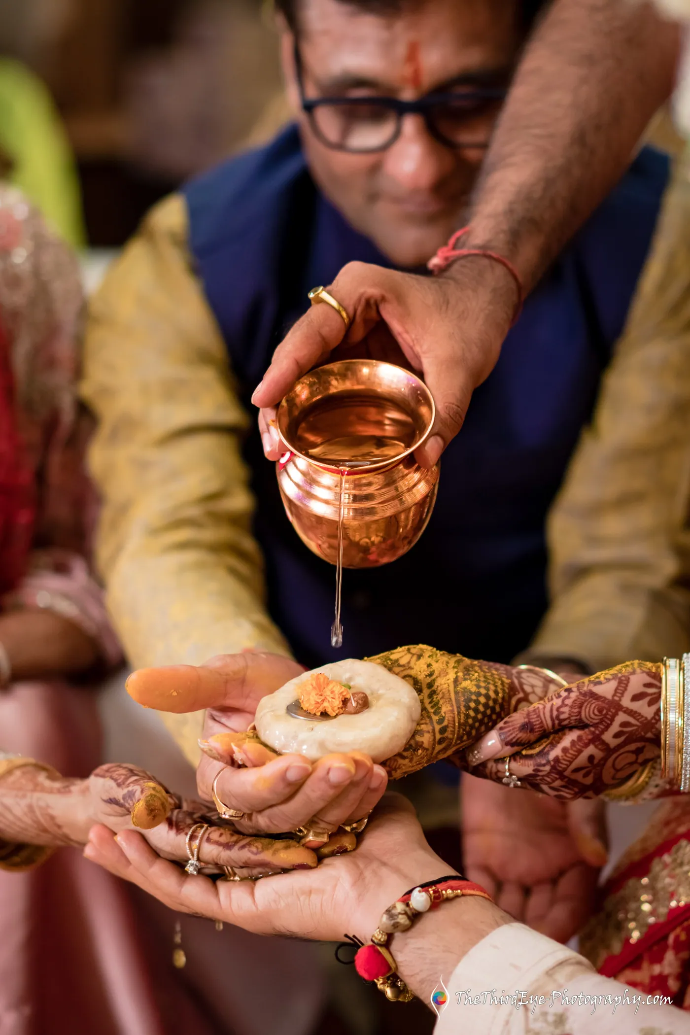 A man in a navy waistcoat tilts a copper pot, pouring water over a stack of hands