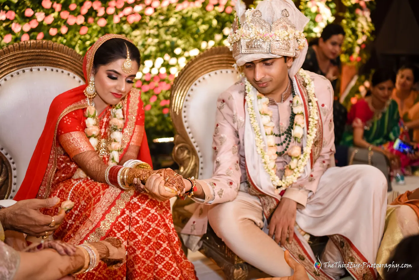 The bride in red and the groom in an ivory sehra sit side by side, hands over the ritual offering