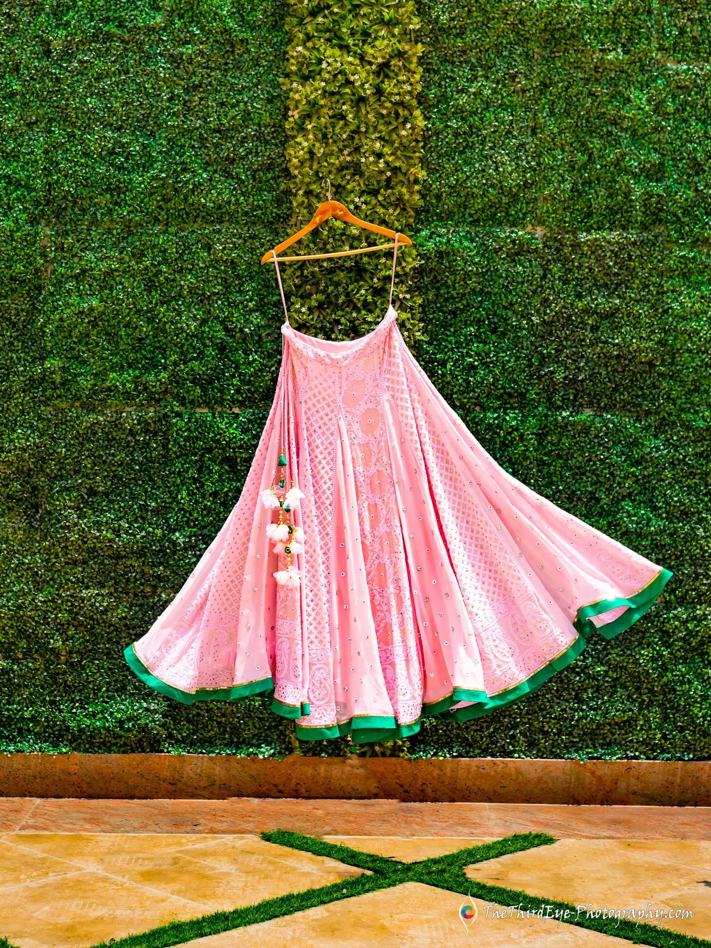 A pink lehenga with a green border hangs on a hanger against a green hedge wall