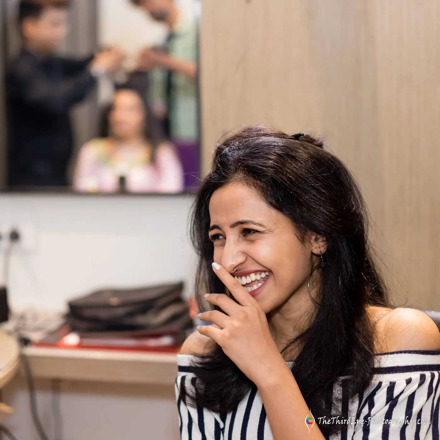 A woman in a striped top laughs behind her hand while a stylist works in the mirror