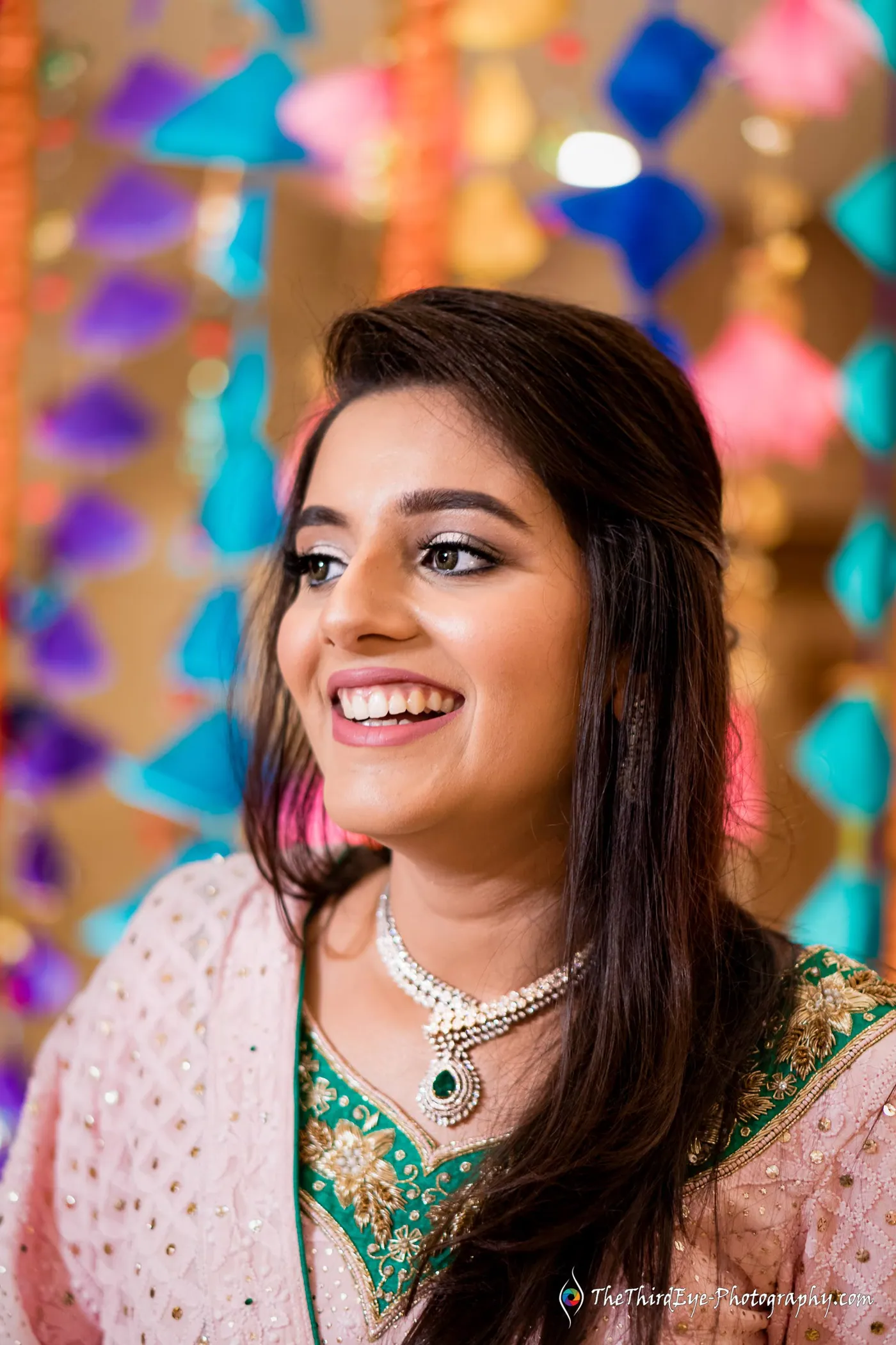 A woman in a pink lehenga smiles wide with colorful paper hangings glowing behind her