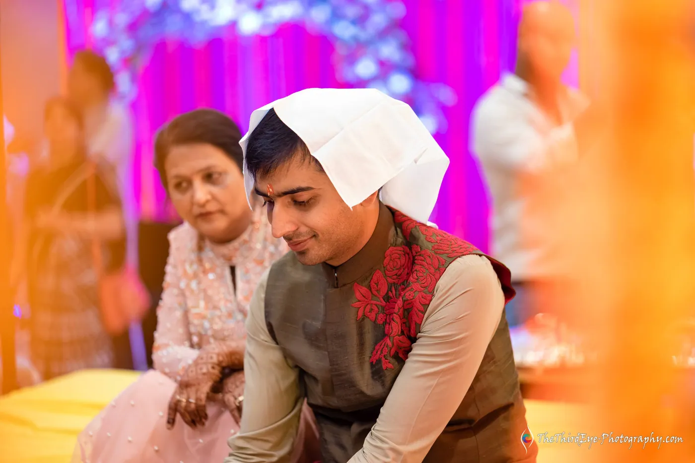A man with a white cloth on his head kneels while an elder woman in a pale outfit watches
