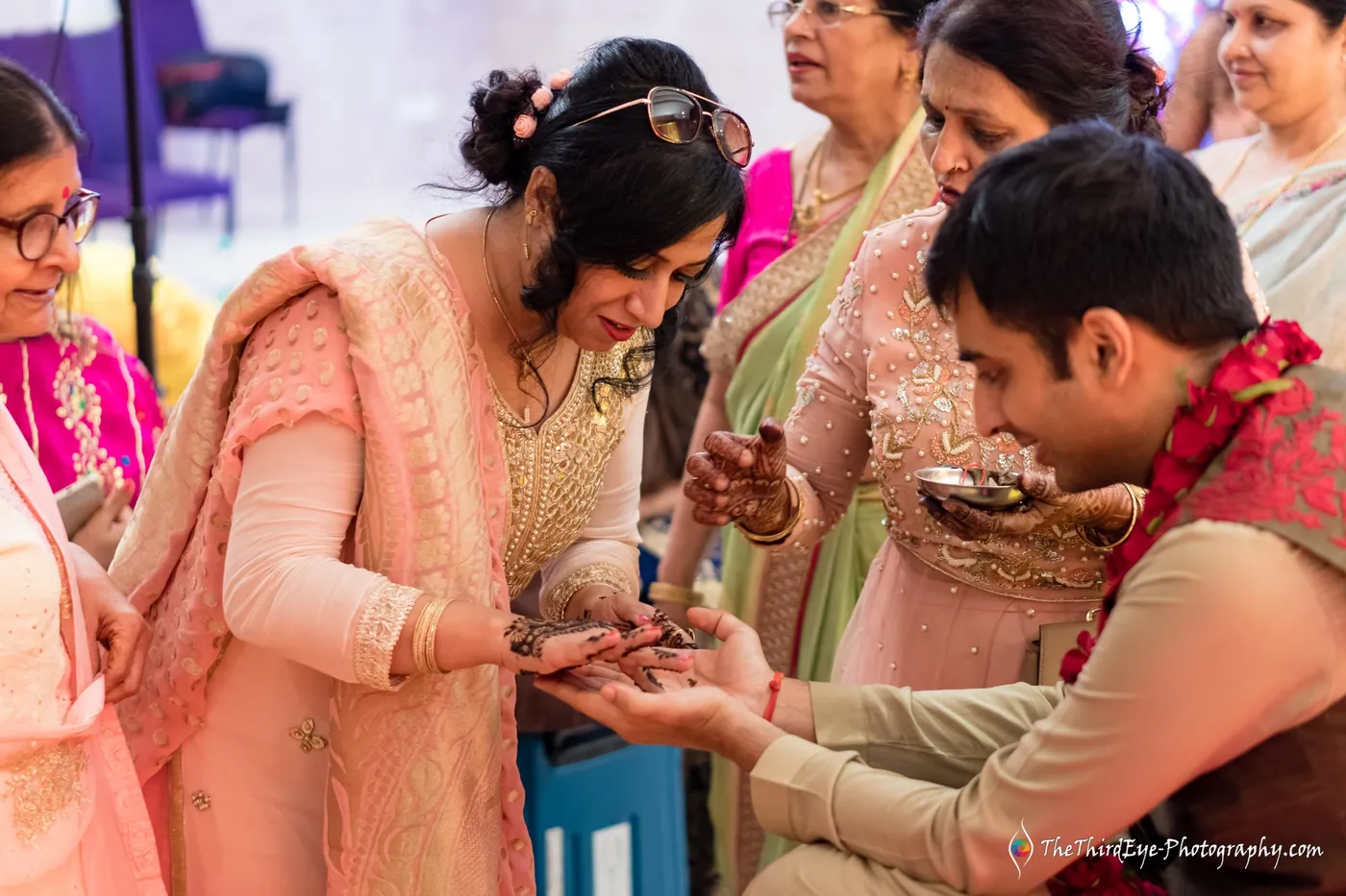 A woman applies henna to the groom's palm as he looks down with a quiet smile, garland on his chest
