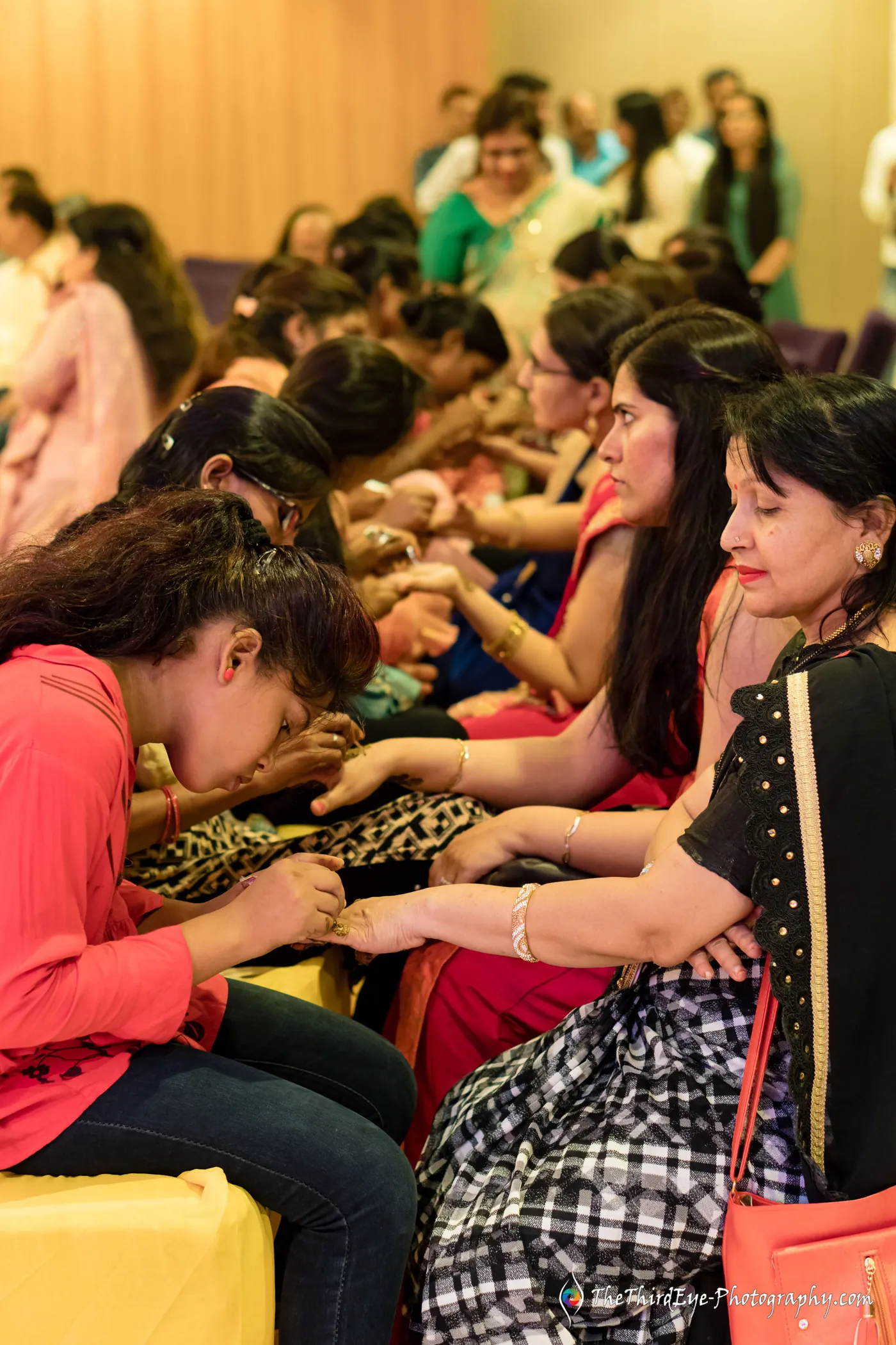 Women sit shoulder to shoulder with heads bent over their hands as henna is applied