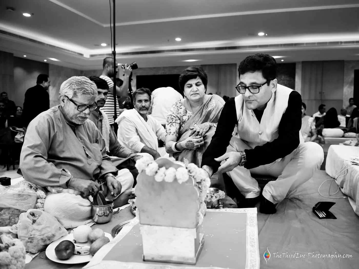 Black and white frame of elders and a young man in glasses gathered around a white block during the pooja