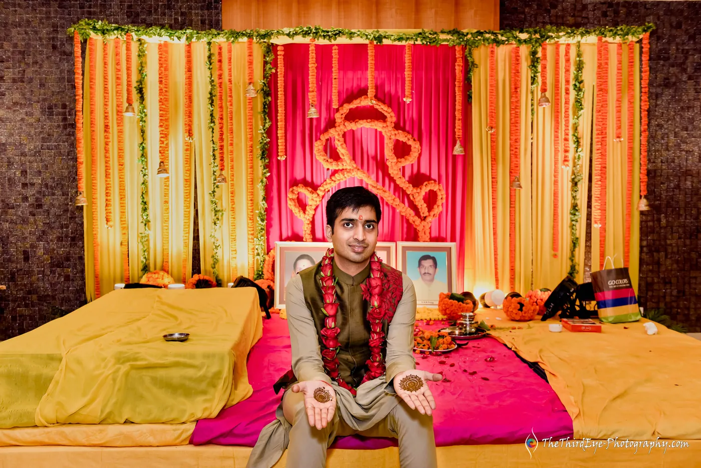 The groom sits on a yellow mattress in a red garland and olive jacket before the pink Ganesha backdrop