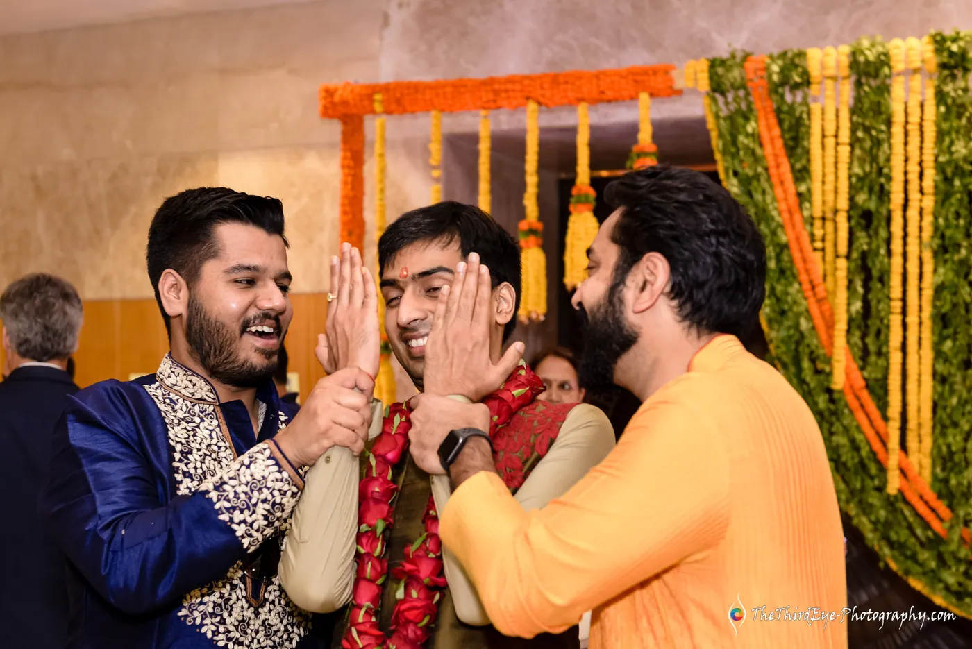 A friend covers a garlanded man's face with a hand while a man in orange leans in laughing