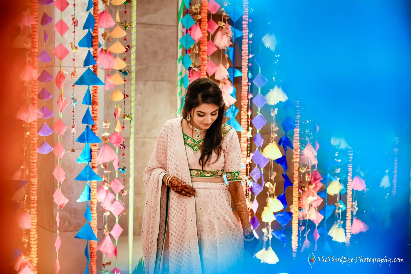 The bride in a pink and green outfit stands among hanging paper kites, looking down at her henna