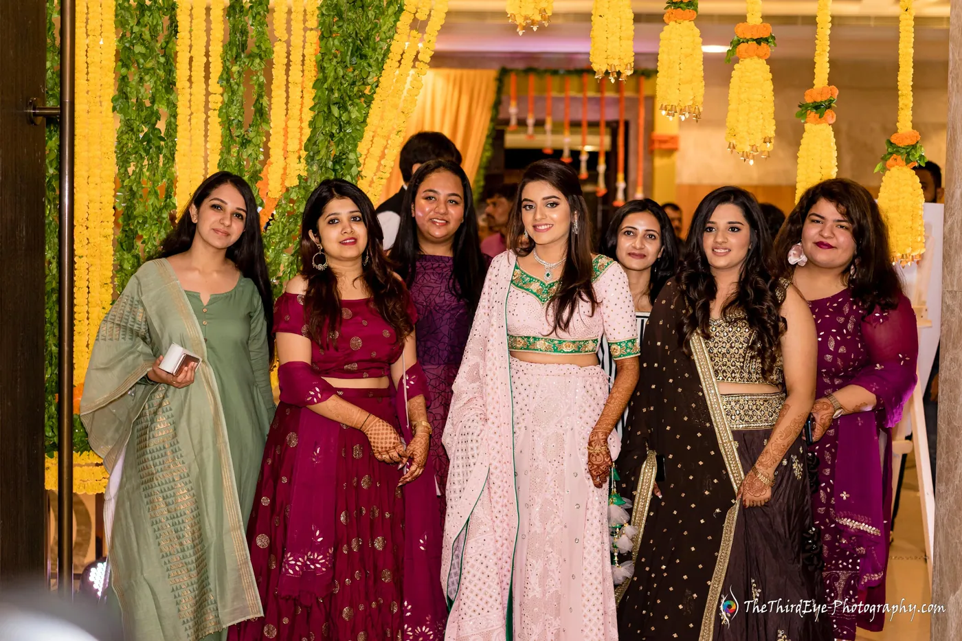 A row of seven women in colourful outfits stand close and laugh in front of yellow garlands