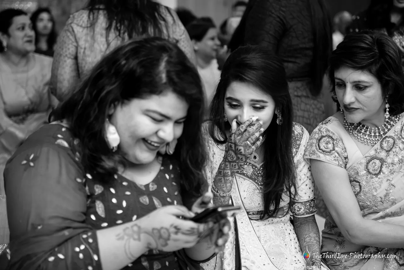 Black and white frame of two women giggling over a phone, one covering her mouth with a henna decorated hand
