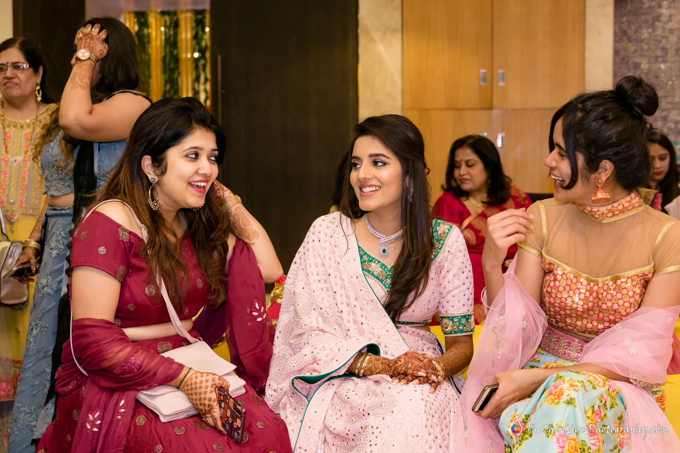 Three women sit and smile together during the mehendi, one in a pink and green outfit