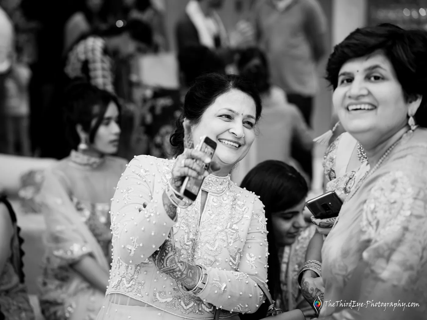 Black and white woman in a sparkling jacket smiles at a raised phone as another woman laughs beside her