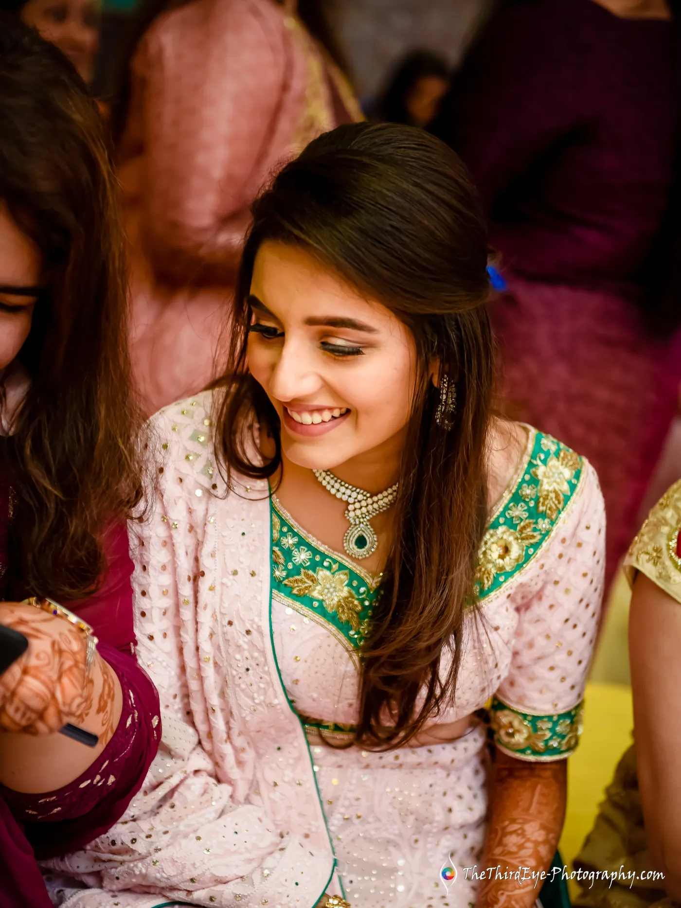 The bride looks down with a wide smile in pink and green, an emerald necklace and henna on her hands