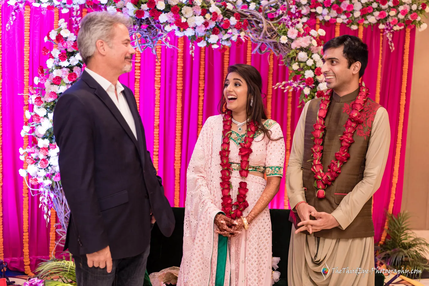 A man in a navy suit stands beside the garlanded couple against a pink curtain as Arushi bursts into laughter