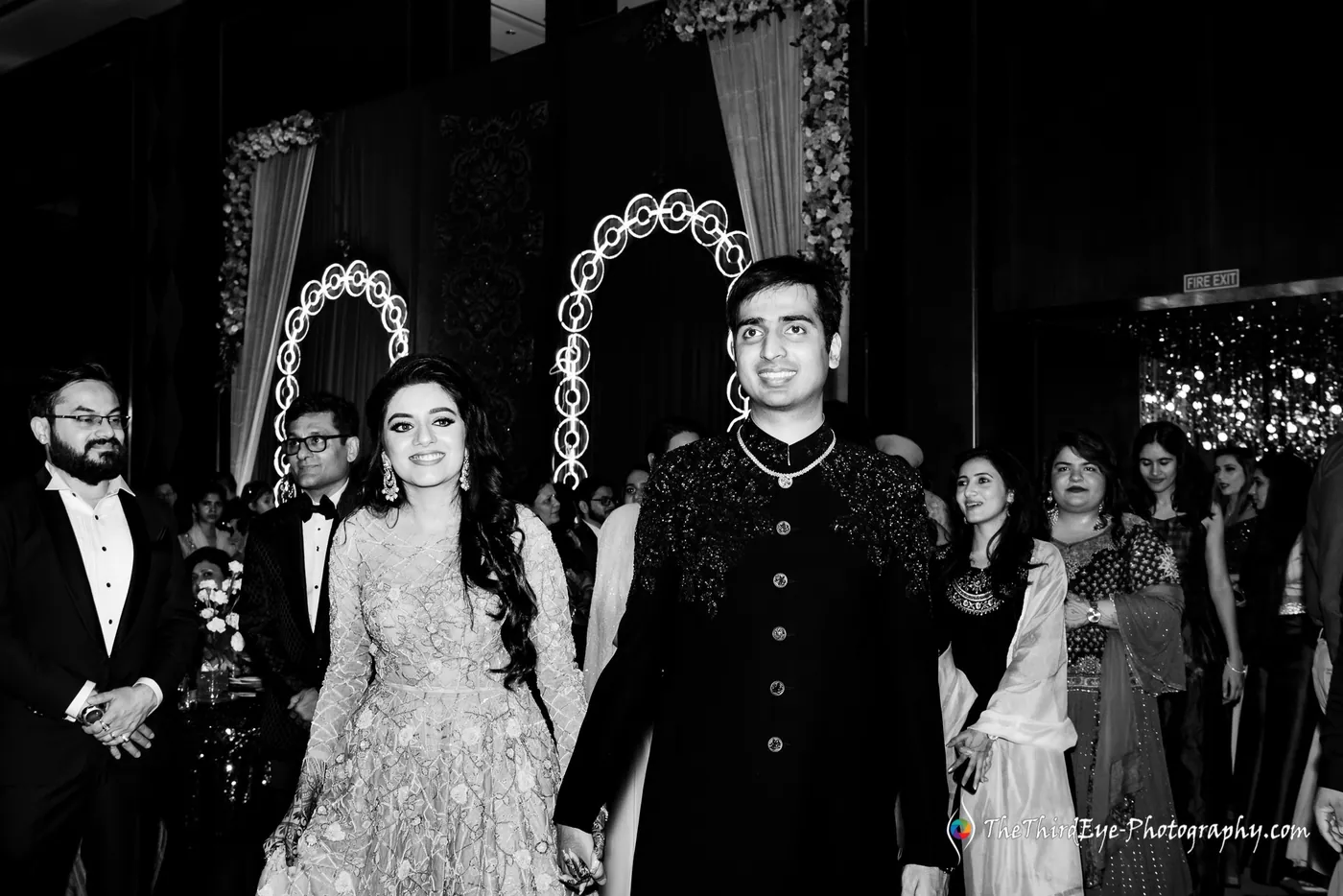 Black and white frame of Ramit and Arushi walking toward the stage in a dark sherwani and a flowing gown