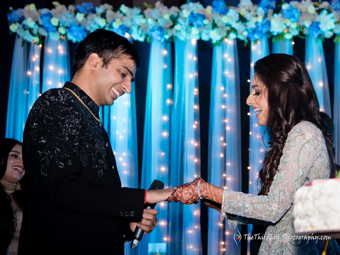 Ramit holds a microphone and Arushi's hand as they stand together under strings of lights