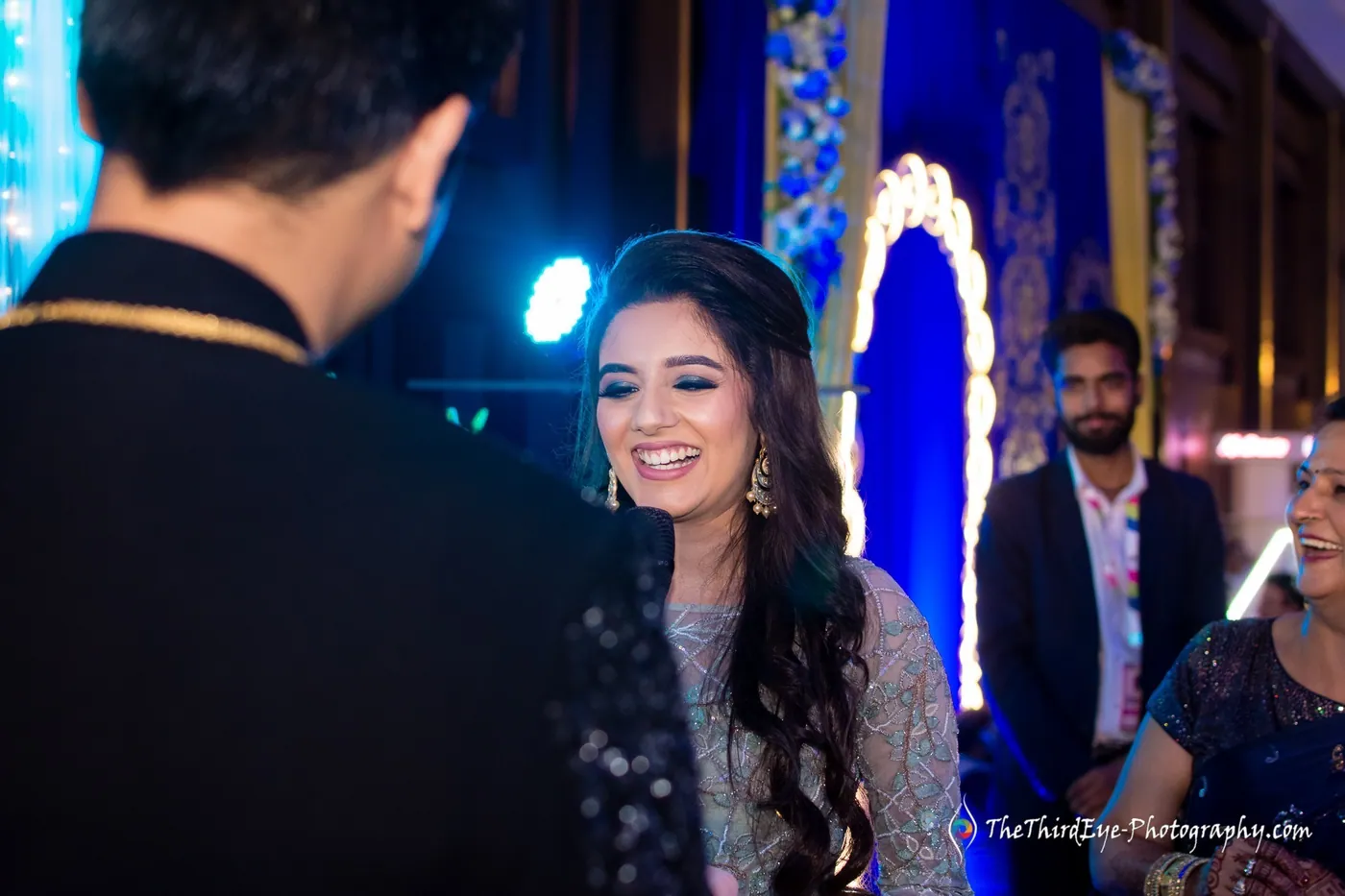 Arushi smiles broadly toward a man in a dark sherwani whose back is to the camera, blue stage lights behind