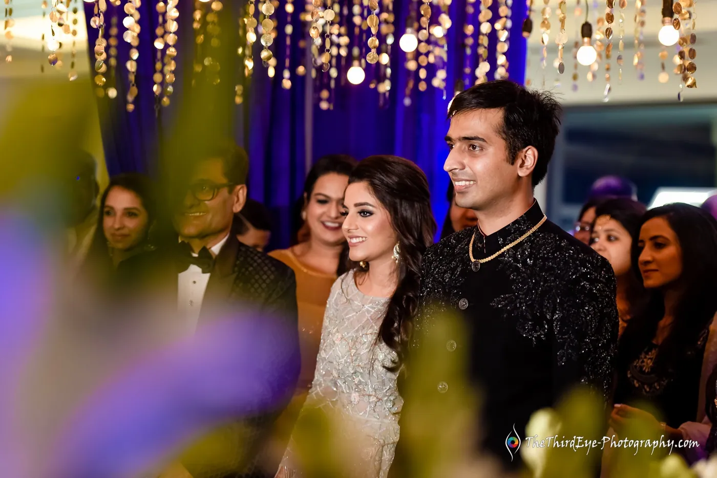 Ramit and Arushi stand side by side among friends who beam at them, leaves blurred in the foreground