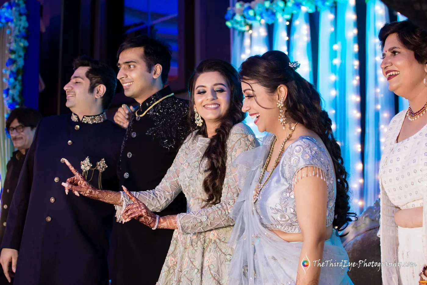 Guests crowd the stage under blue lights, one in a pale gown laughing and a man pointing out of frame
