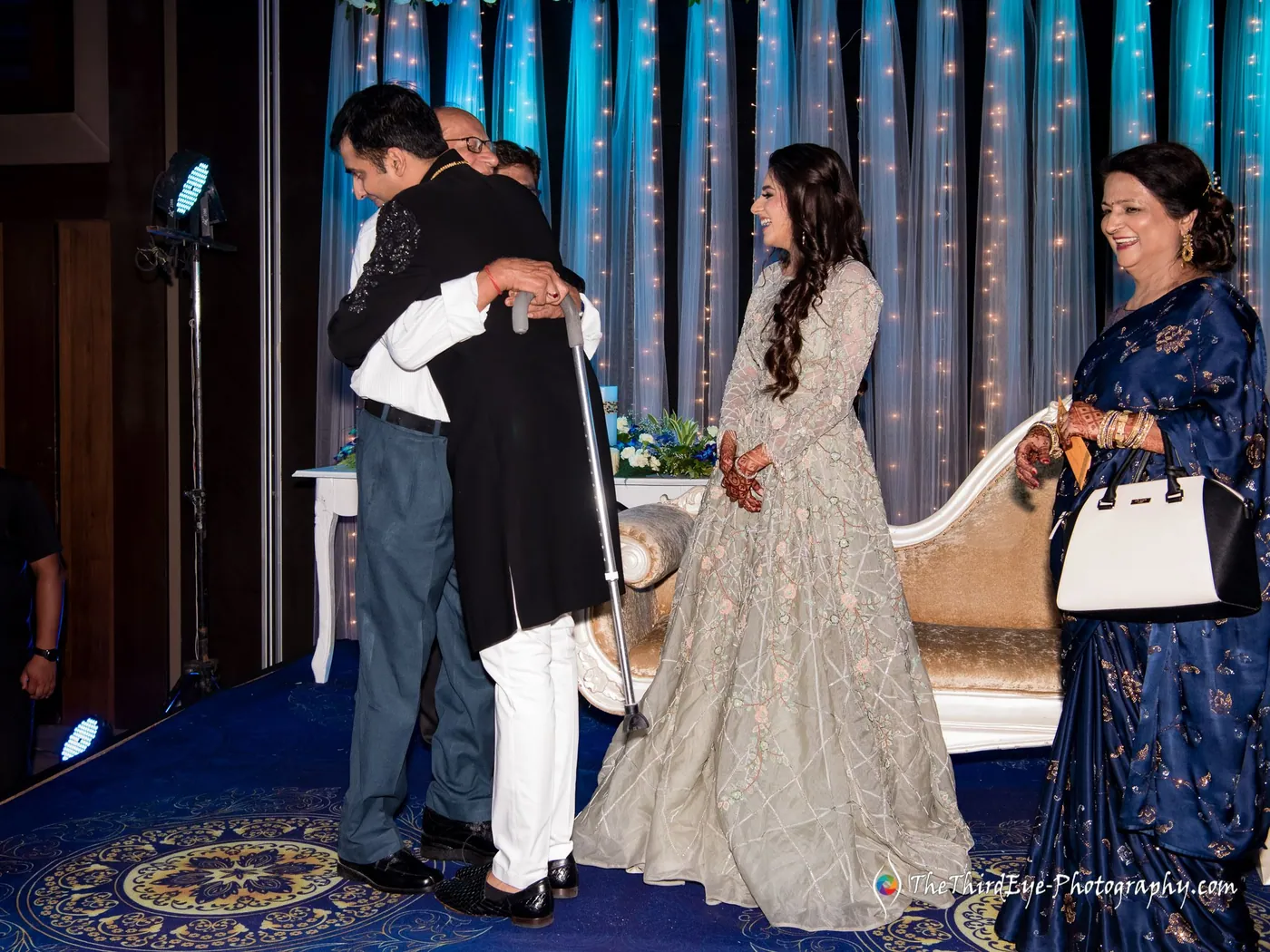 Two men share a hug as Arushi in a pale gown waits with a woman who holds a handbag