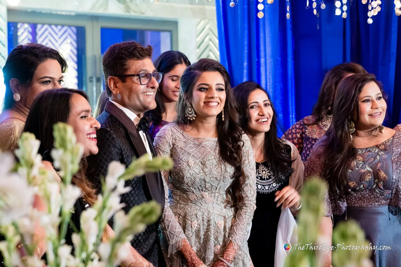 Arushi stands among relatives who laugh and smile, white flowers blurred in the foreground