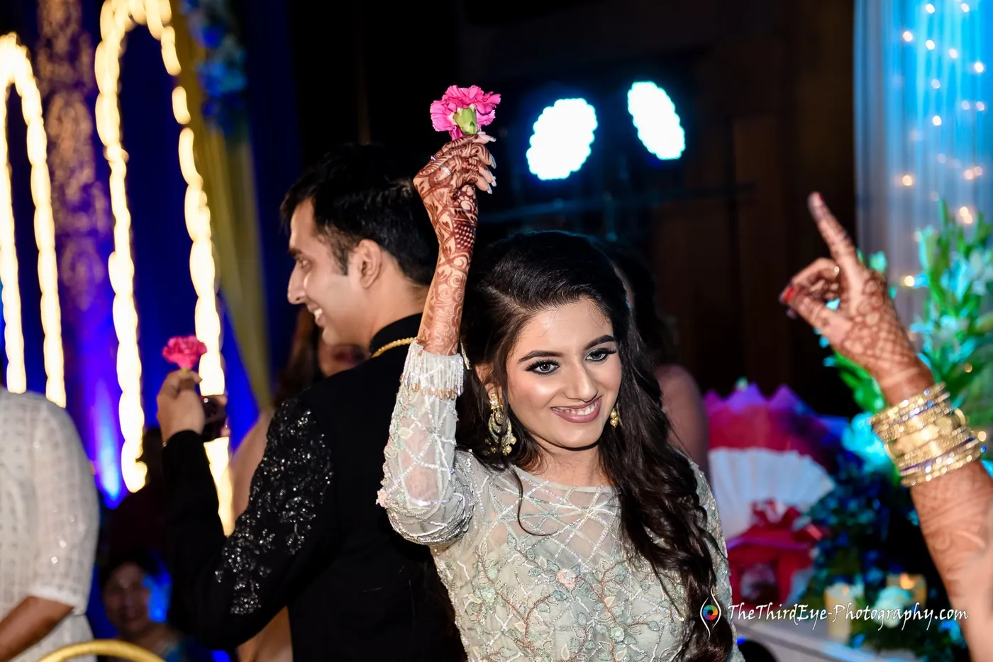 Arushi holds a pink flower and points a finger skyward, laughing, as Ramit stands behind her in a dark jacket
