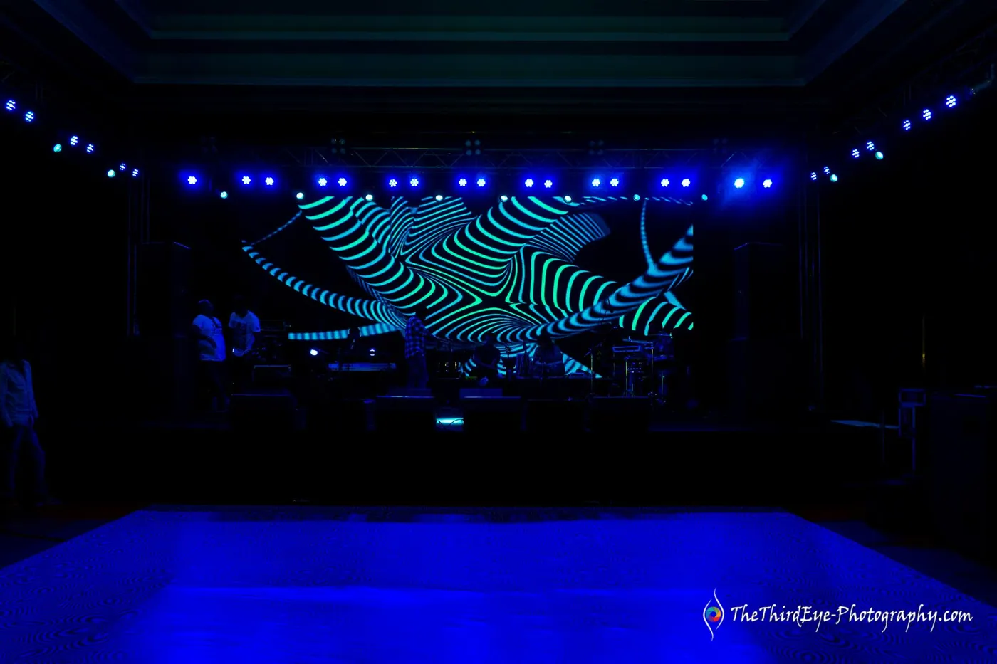 A dark ballroom with a blue floor, dotted lights and an empty stage glowing in green waves