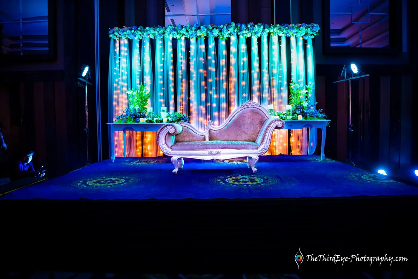 A cream settee sits before a teal and gold flower backdrop on a blue lit stage