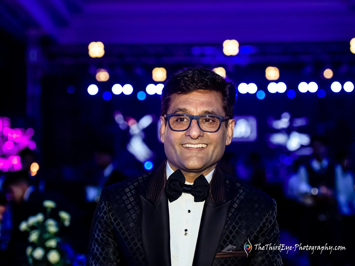 A man in a tuxedo and bow tie grins at the camera under blue party lights