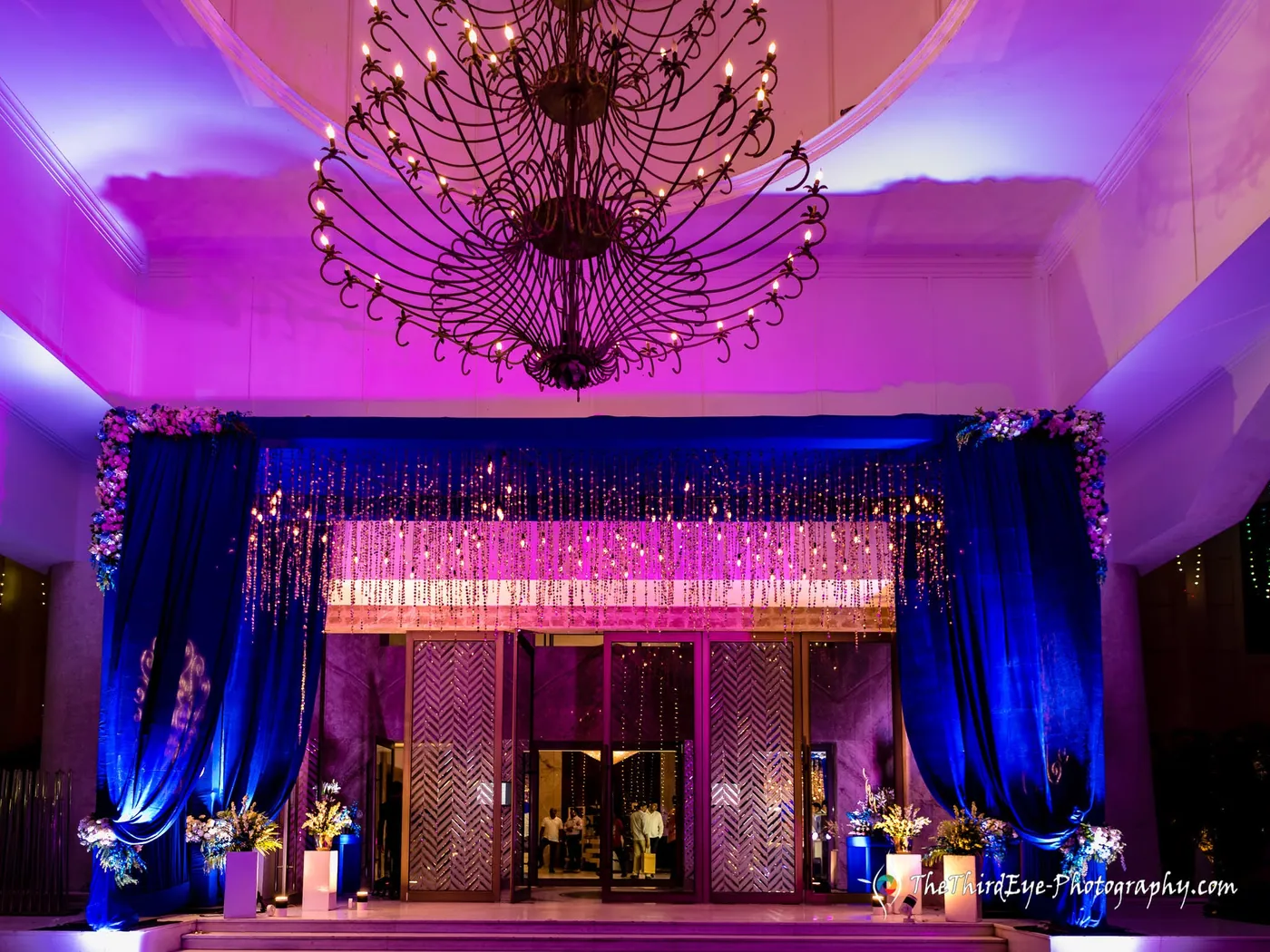 Grand ballroom entrance under a crystal chandelier, deep blue drapes framing a pink glowing doorway