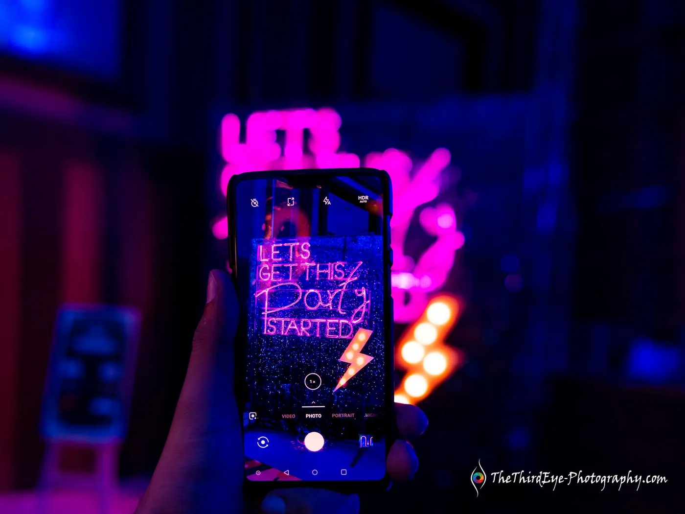 A phone held up to film a pink neon sign that reads Lets Get This Party Started