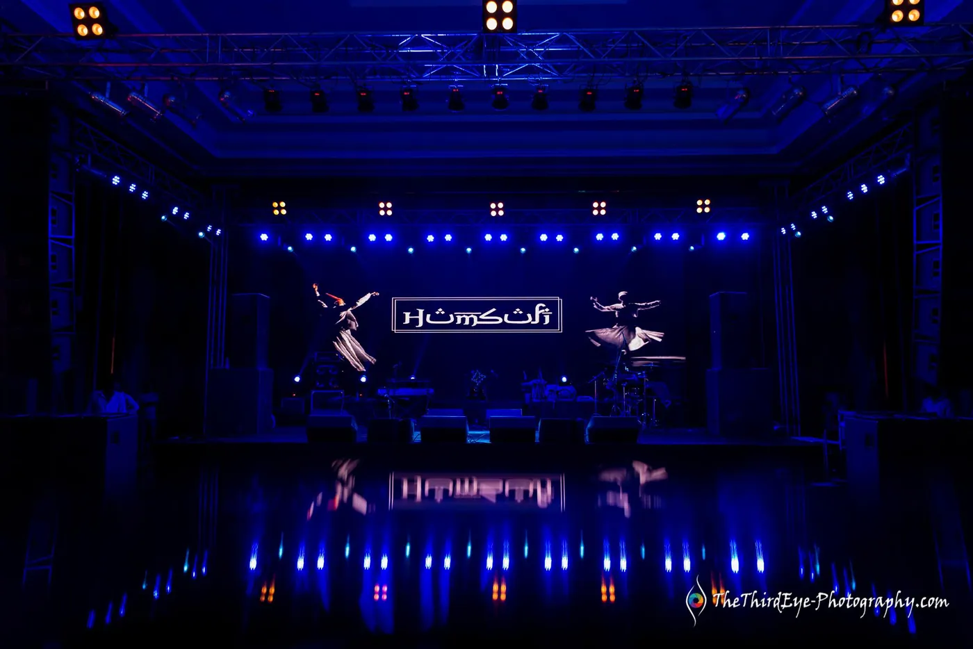 Wide view of the stage under blue truss lights with the Humsufi logo above the band
