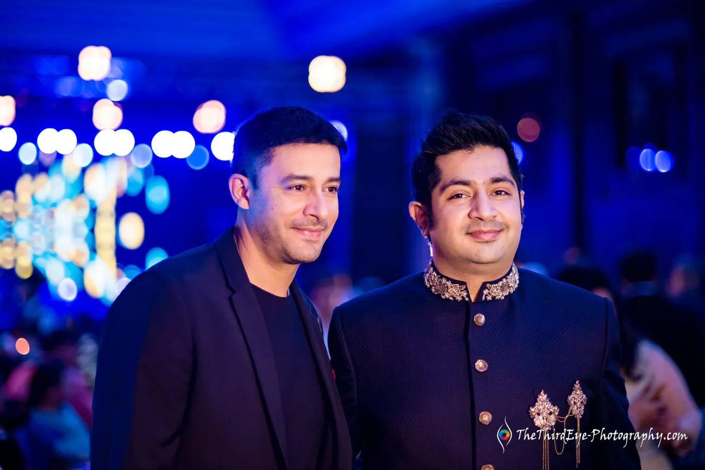 Two men in dark outfits stand close and smile at the camera against blurred festival lights