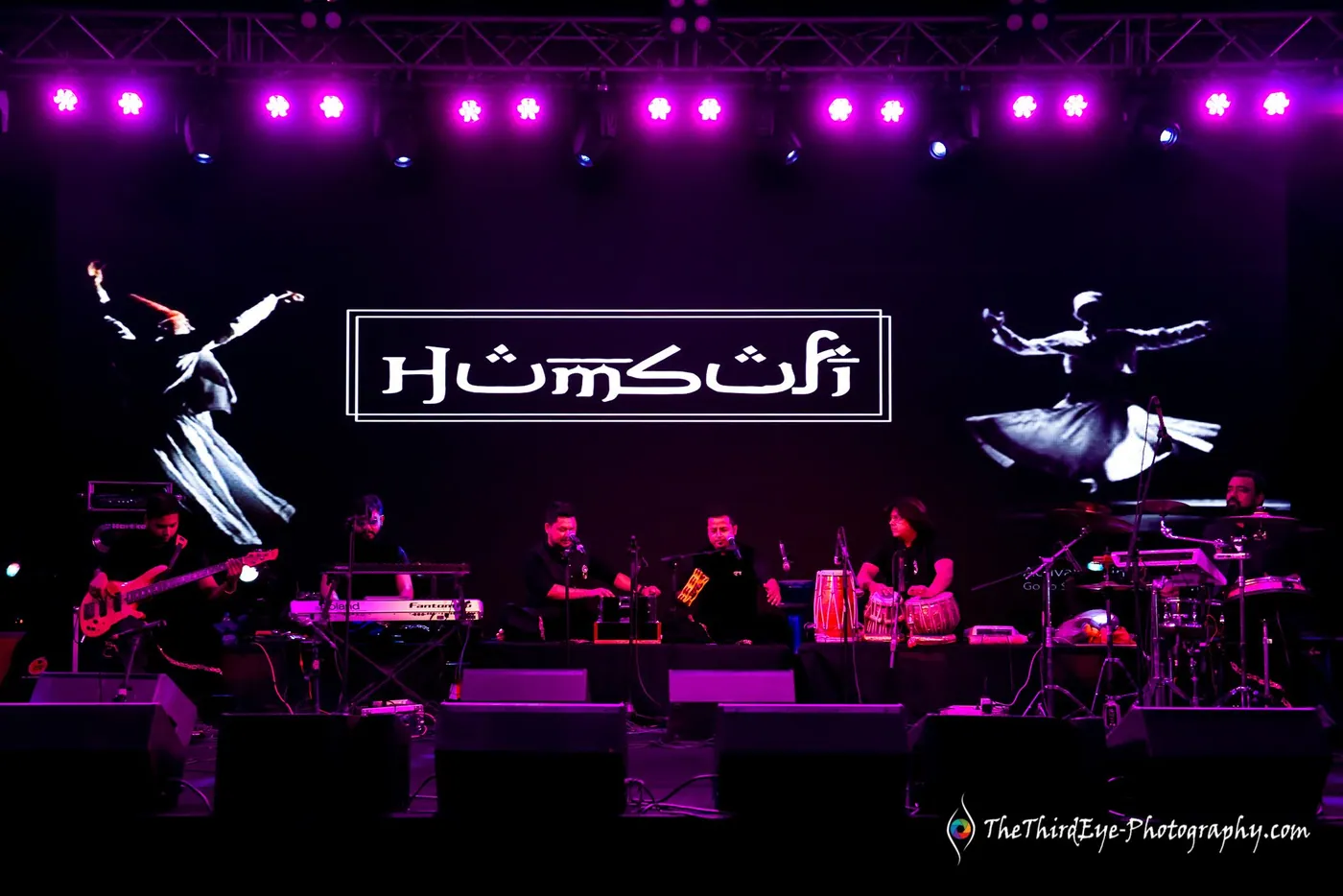 The Humsufi logo glows above the band on a purple lit stage, dancing dervish figures on the backdrop