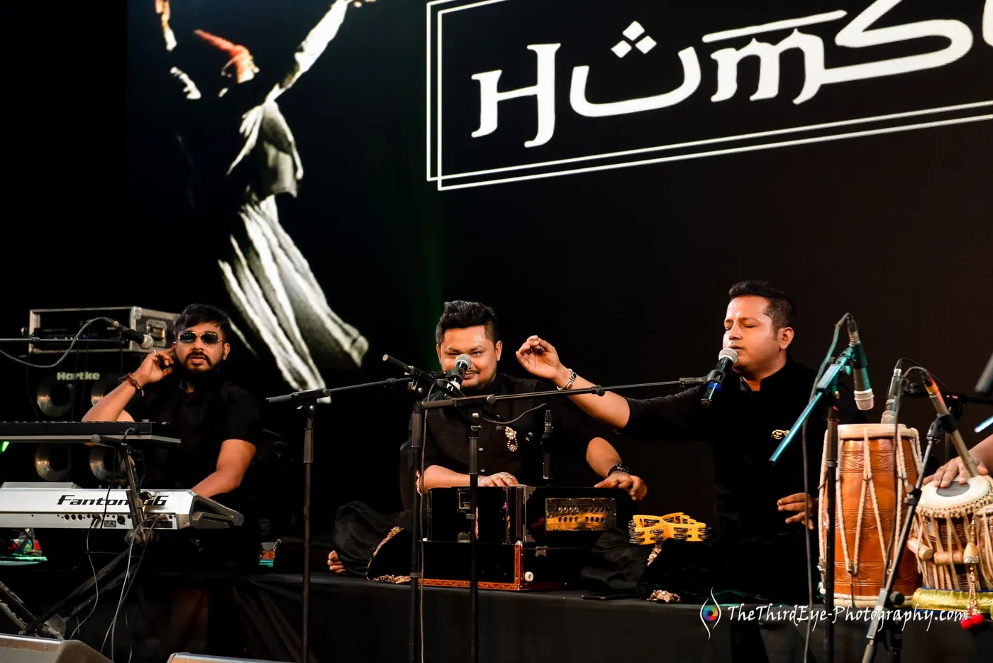 Humsufi musicians play keys and dhol beneath the lit group logo with a whirling dervish graphic