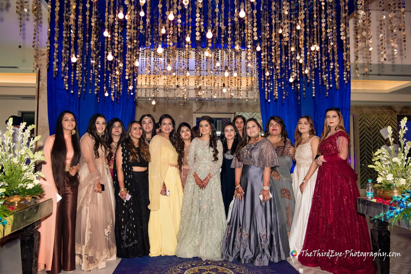 A long line of women in gowns and lehengas pose together before a glittering curtain