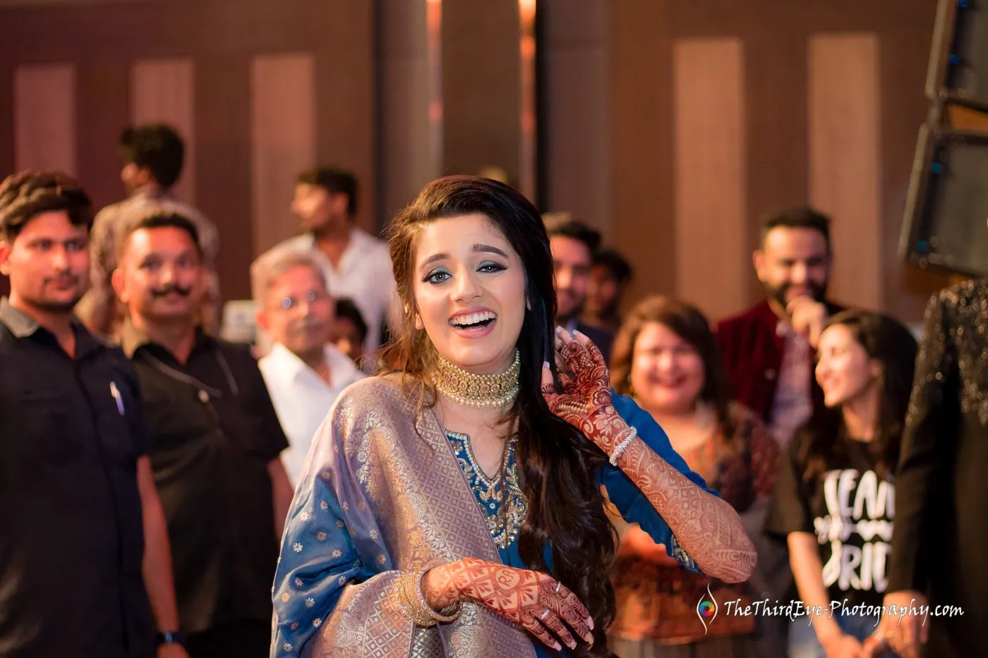 A woman with henna decorated hands laughs wide in warm light as a crowd smiles behind her