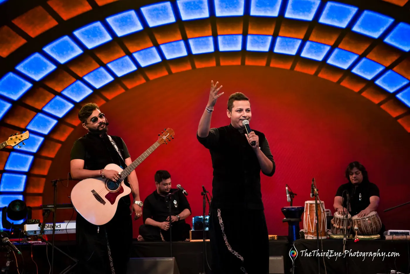 The lead singer raises a hand mid song as a guitarist and tabla player flank him