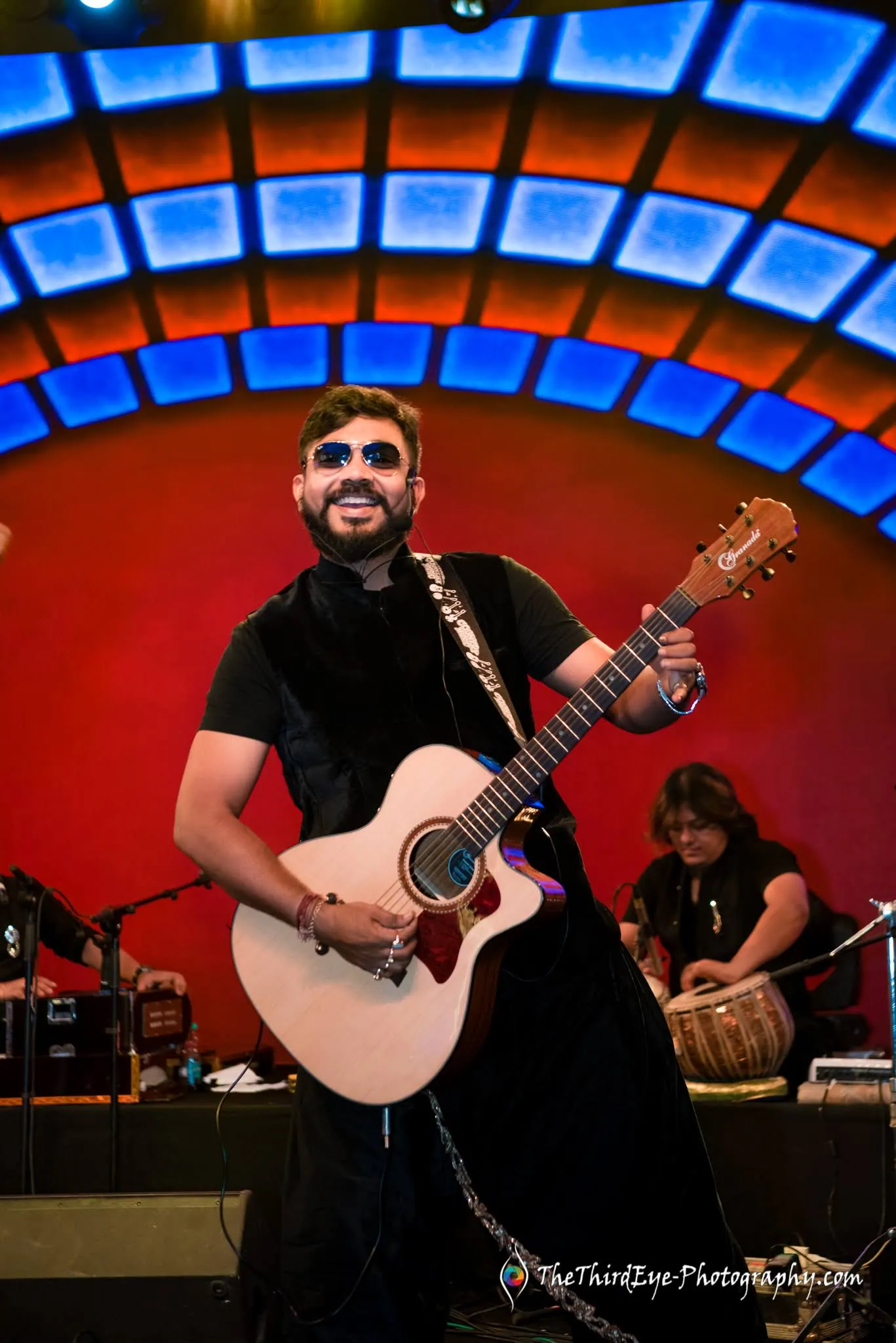 A guitarist in sunglasses grins on the red arched stage with a tabla player behind him