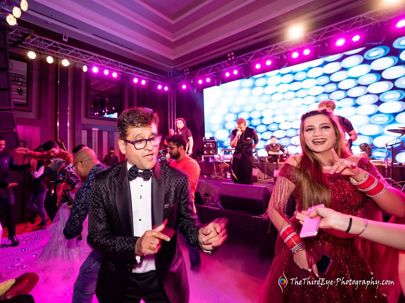 A man in a tuxedo and a woman in red dance with big smiles under pink stage lights