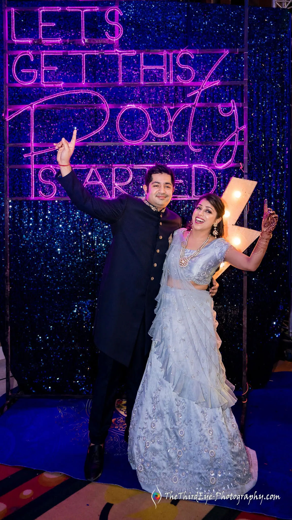 A man in a dark sherwani and a woman in pale blue raise their hands before a neon sign