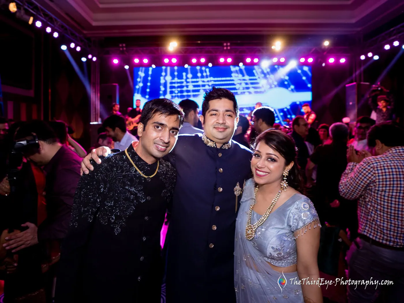 Two men in dark sherwanis and a woman in a pale sari squeeze together before the lit stage and crowd