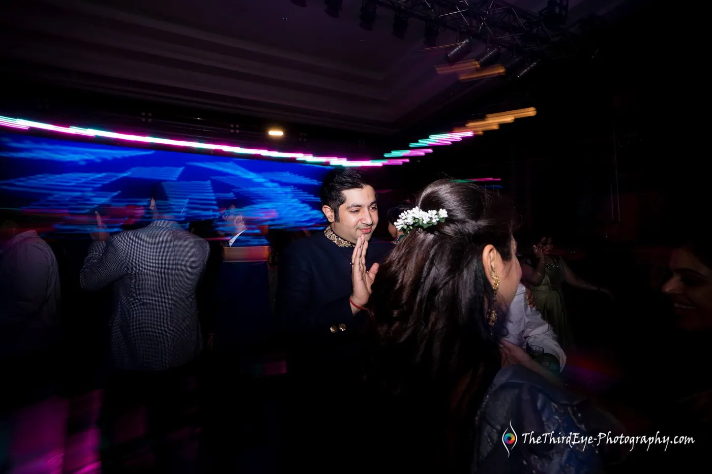 A man in a dark sherwani turns toward a woman with flowers in her hair as blue light streaks