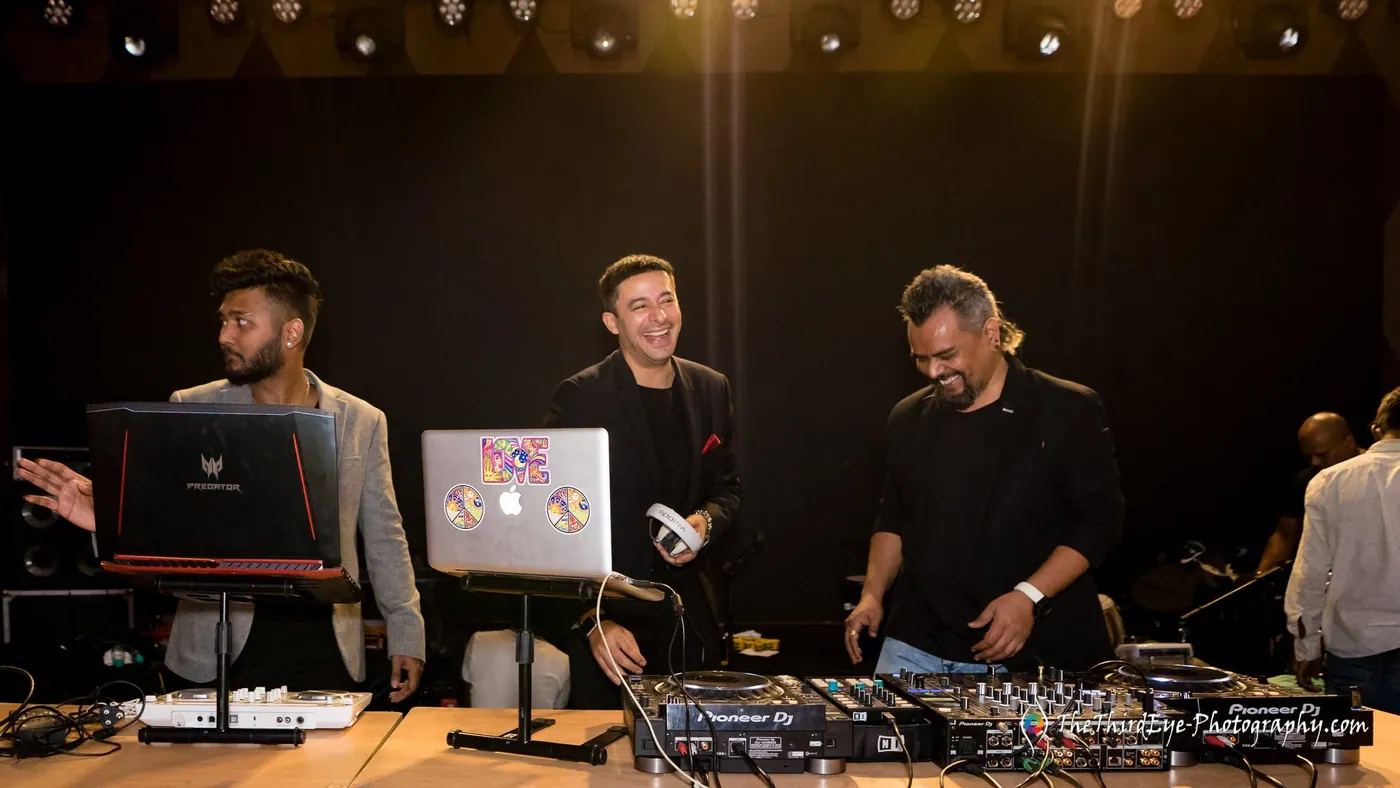 Three men stand behind DJ decks and laptops, the one in the middle laughing