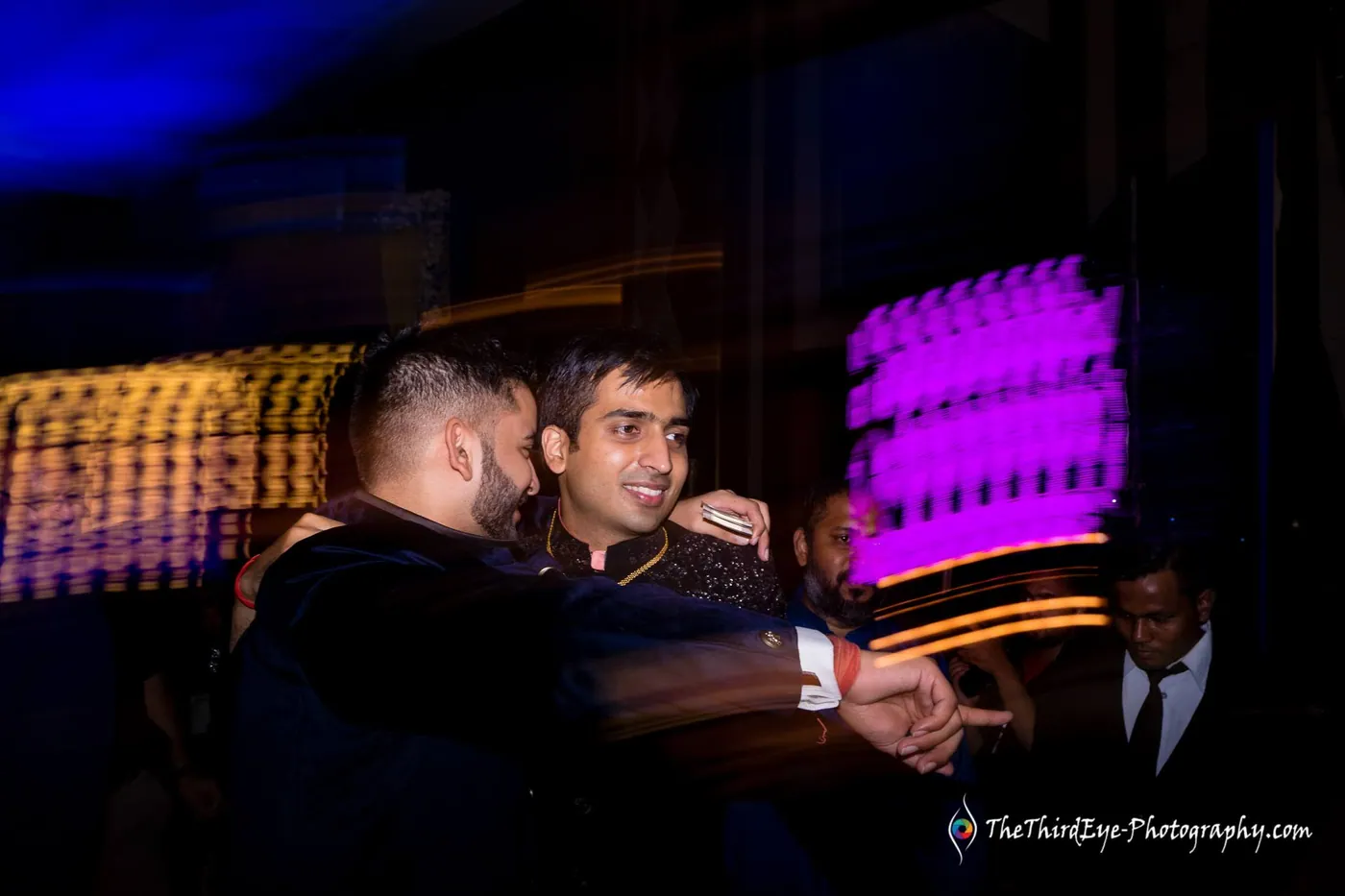 Slow shutter blur around two men holding each other close in a dark hall with a purple screen behind