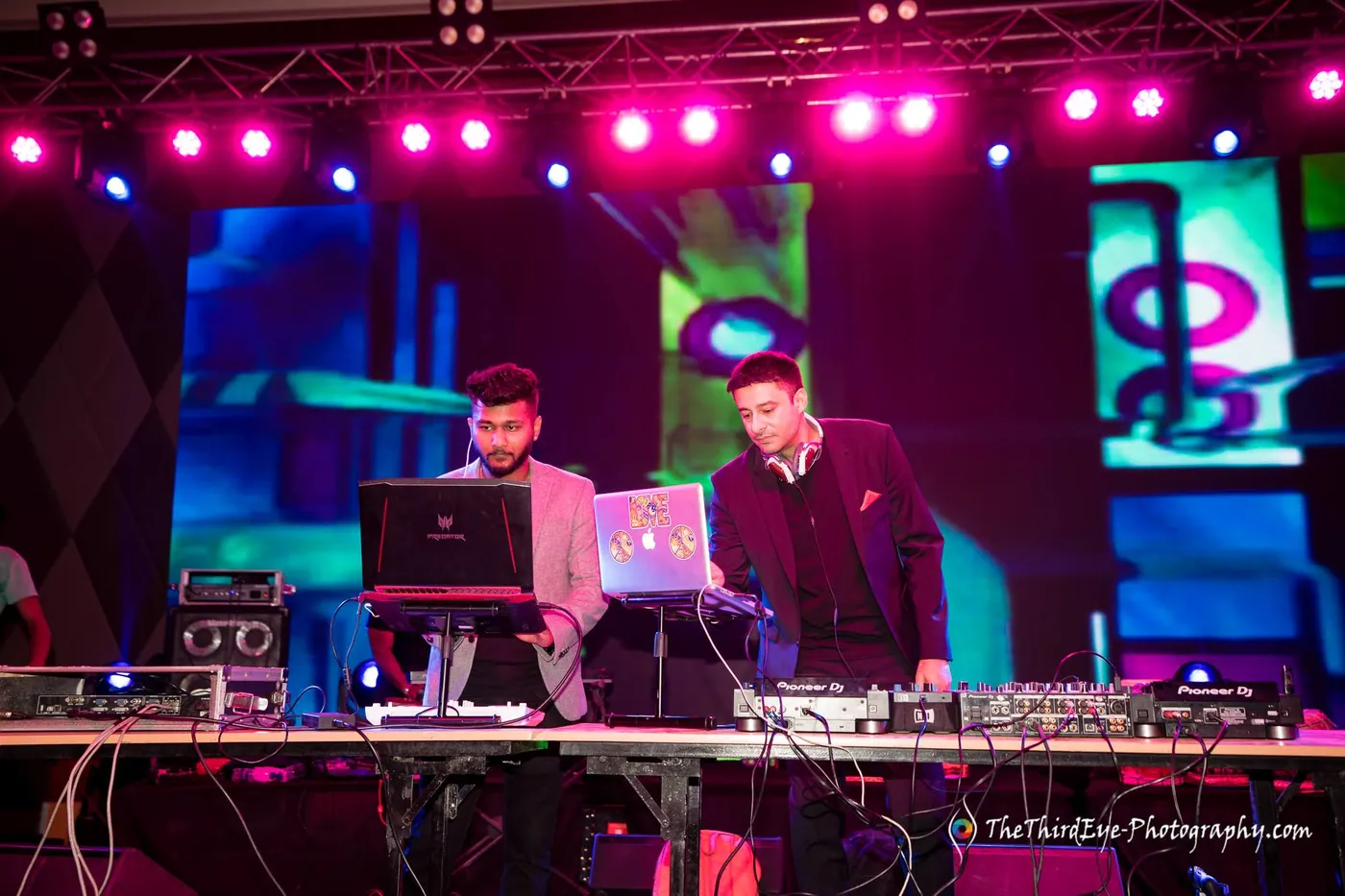 Two DJs work the decks and laptops on a lit stage with a pink truss rig above them