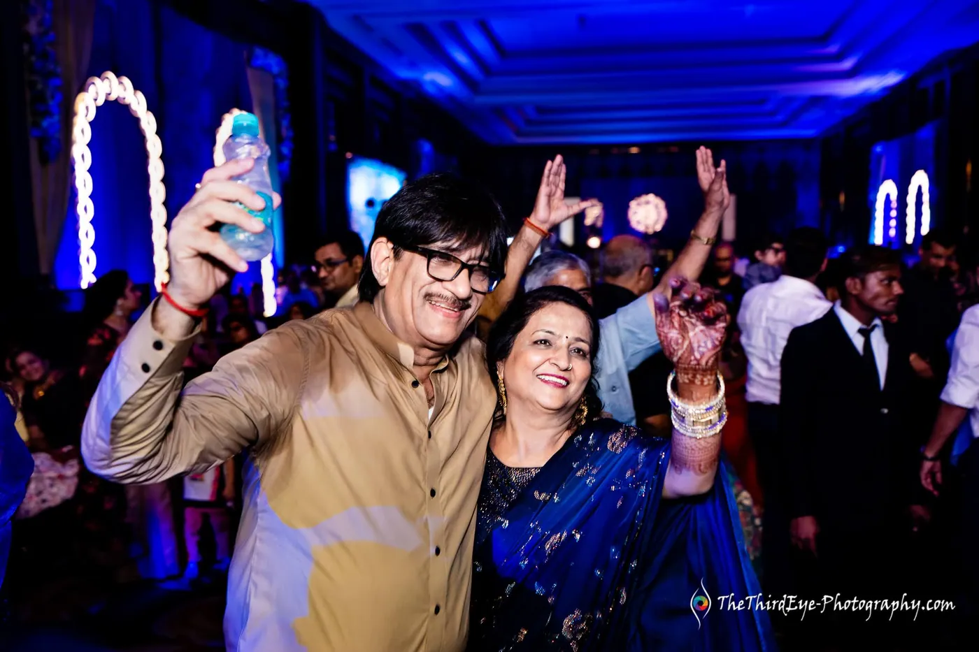 An older man in a gold kurta and a woman in blue dance with raised arms, a phone held high