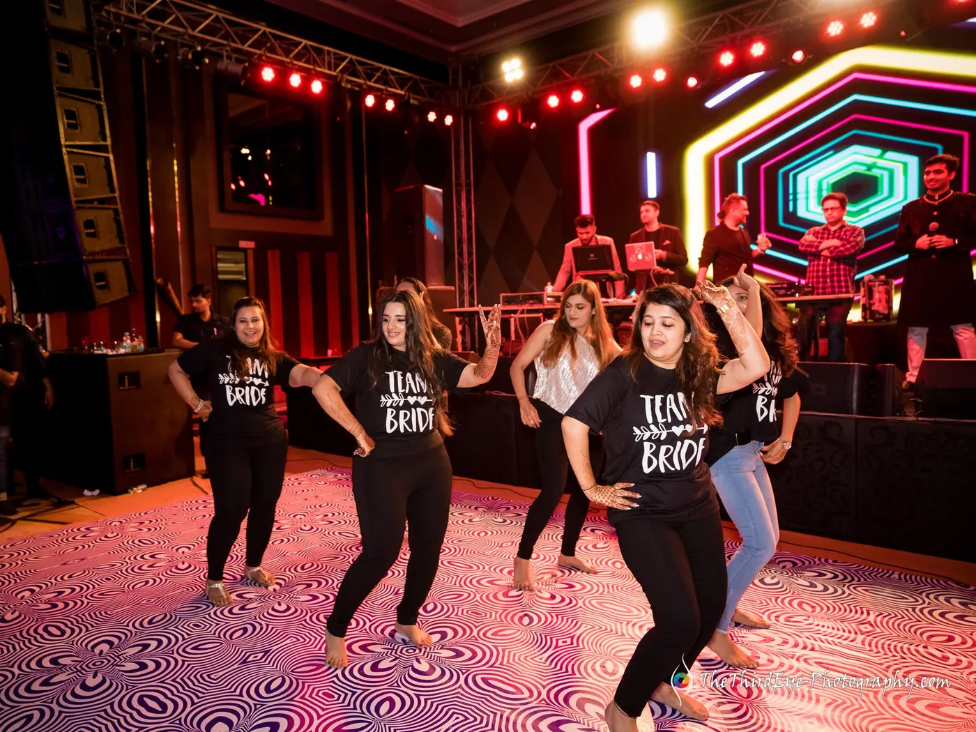 Women in matching black tees dance a routine on a patterned floor under red stage lights