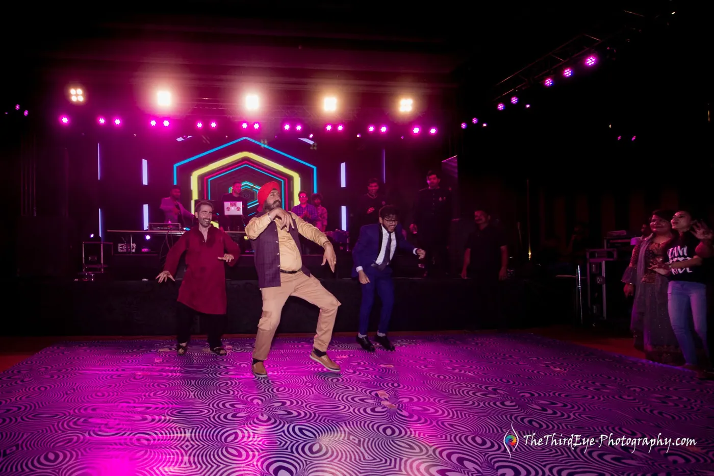 A dance act on the lit stage with a hexagon neon arch and pink beams over a patterned floor