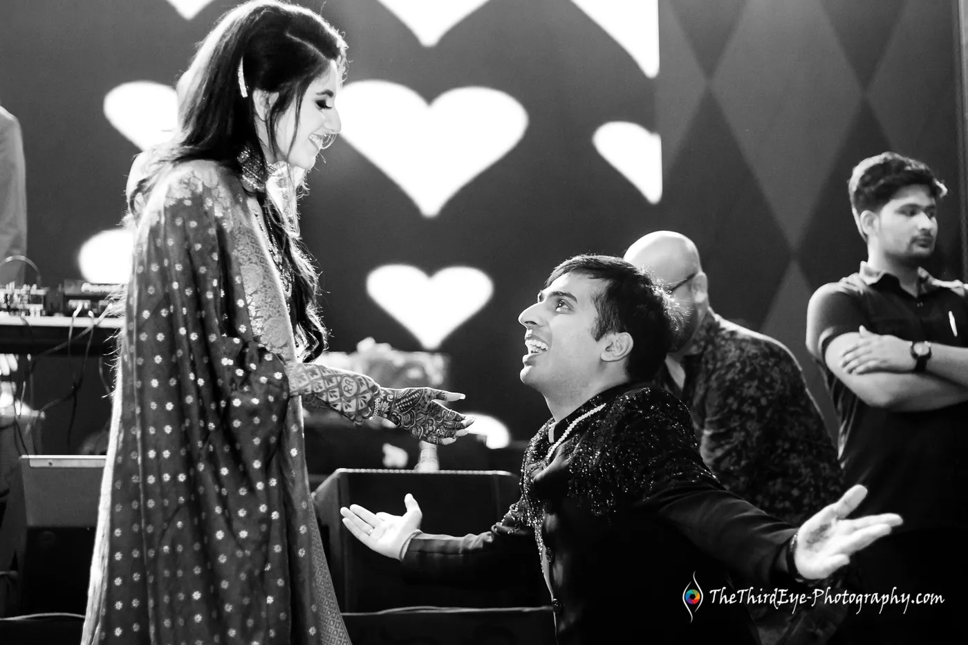 Black and white man on one knee with arms spread before a woman, glowing hearts on the screen behind